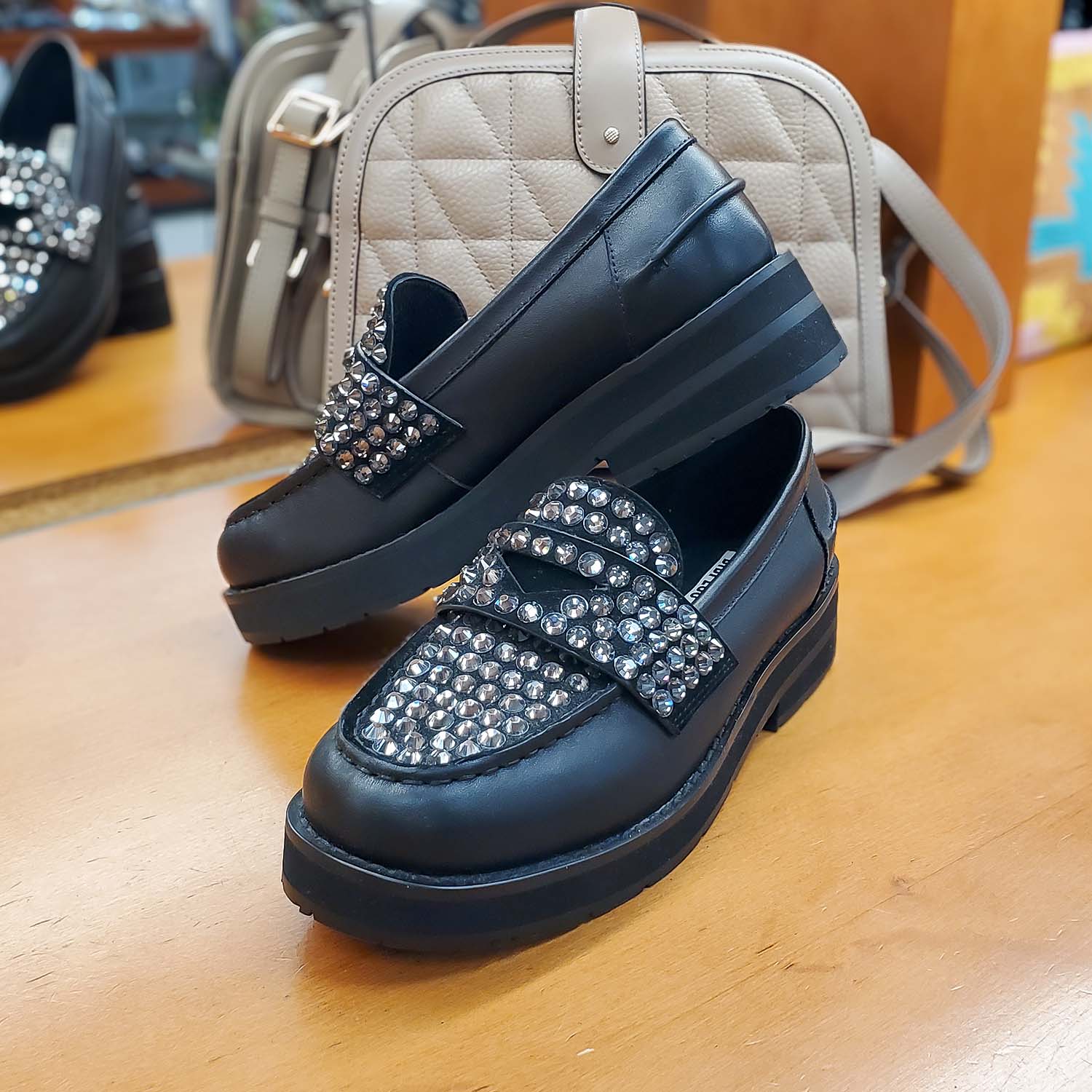 Para czarnych loafersów Bibi Lou z cyrkoniami, ustawiona na drewnianym blacie, w tle torebki
