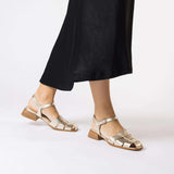 Złote sandały Wonders z paskami na stopach modelki, niski obcas i klasyczna forma w stylizacji z czarną sukienką